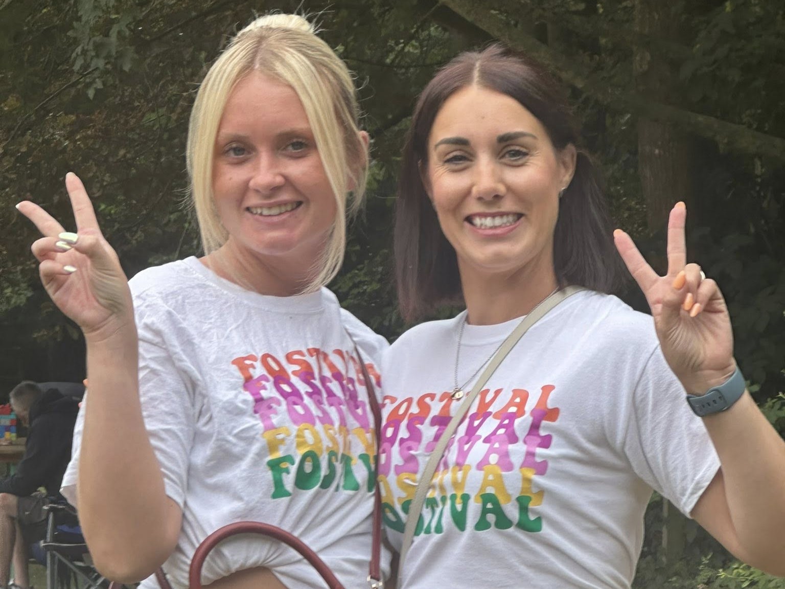 Two women smiling and giving peace signs to the camera. They are both wearing white tops that read 'Fostival'