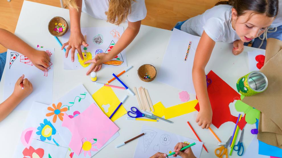 A bird's eye view of a table, with three children making collages using coloured paper 