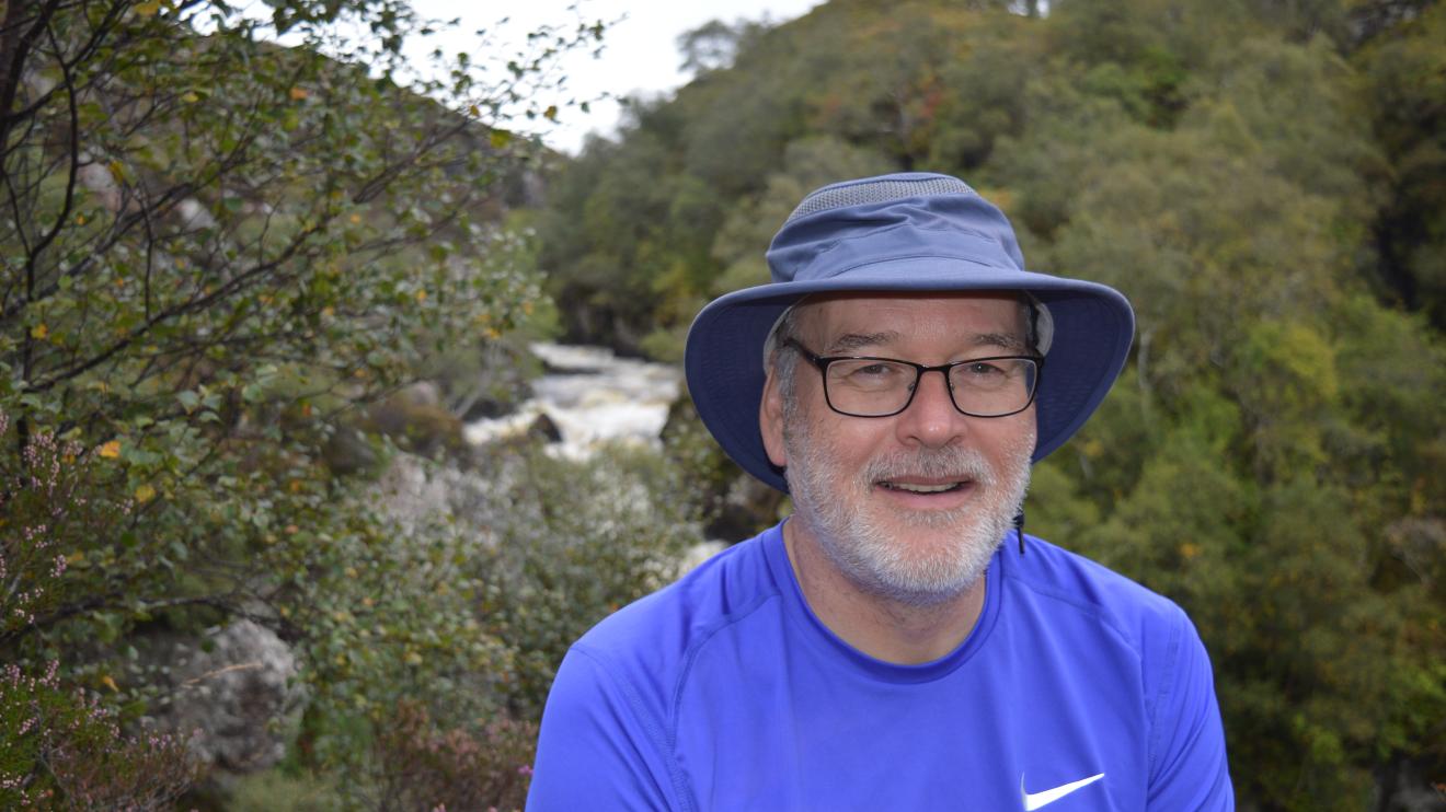 A man sat in front of green trees and foliage. He is wearing a blue hat and is smiling at the camera.