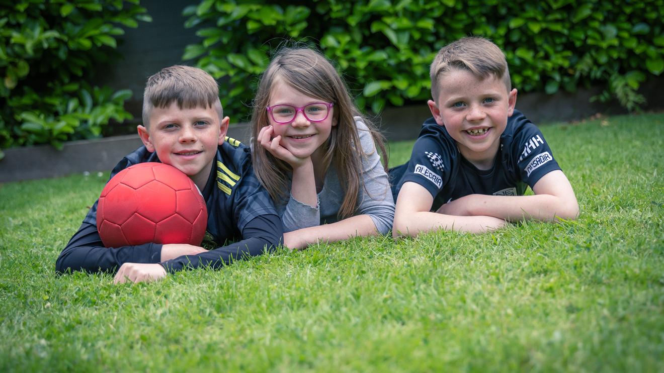 3 children on a grass lawn smiling, one with a red football