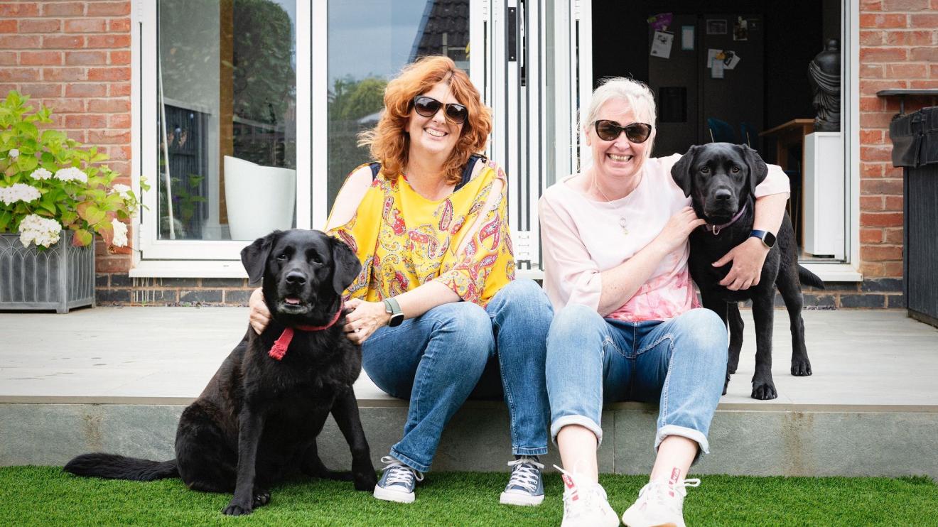 LGBTQ+ foster carers Two women sitting in their garden, smiling, with two black dogs sitting besides them