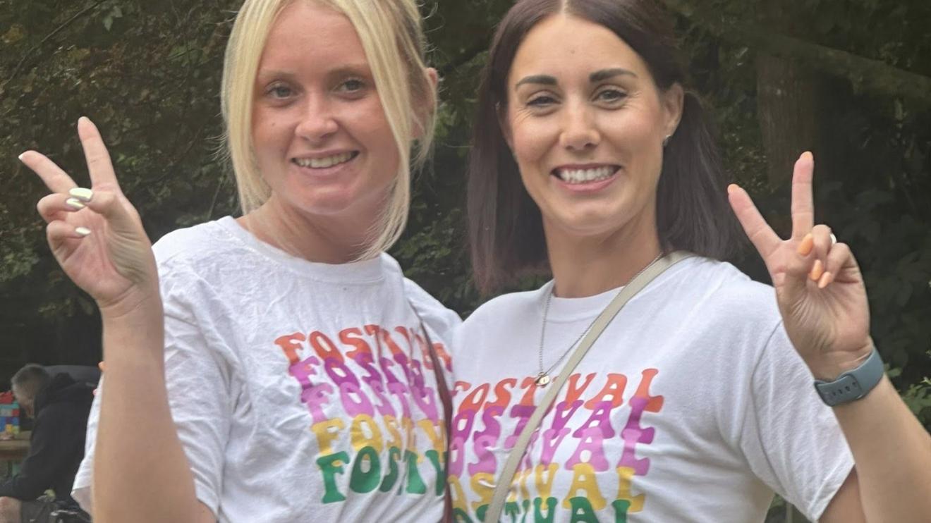 Fostival 2025 - a warm welcome Two women smiling and giving peace signs to the camera. They are both wearing white tops that read 'Fostival'