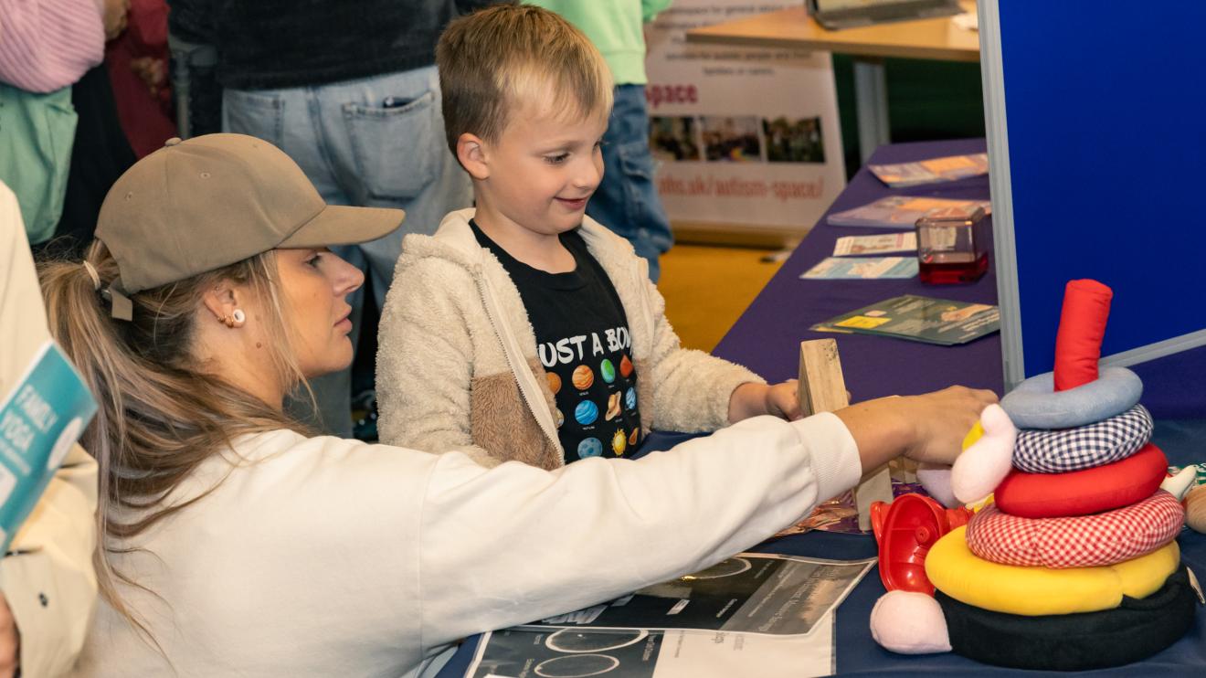 Woman with white jumper and a cap pointing to to a colourful stacking ring toy while a small boy watches on