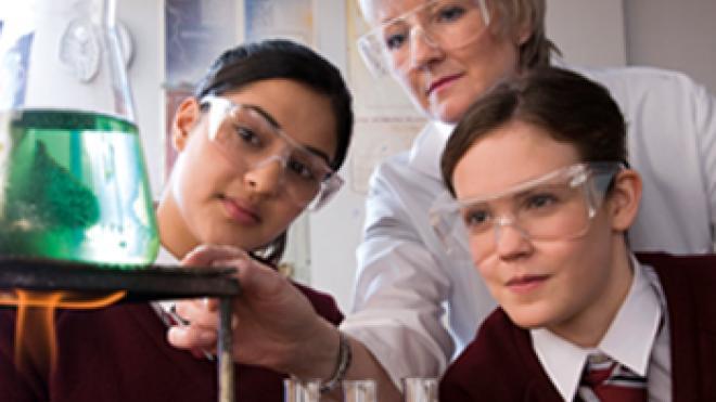 Two secondary school children and a teacher, wearing goggles and looking at scientific equipment.